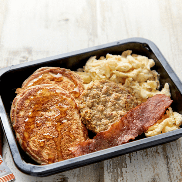 Whey & pea protein pancakes topped with maple syrup, chicken sausage, chicken bacon, and egg scramble served in a dishwasher-safe container