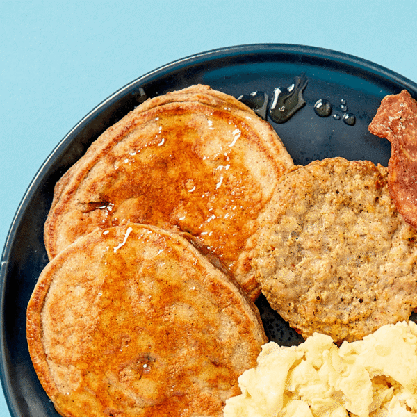 A close-up of a nutritious plate from Fitlife Foods containing two pancakes topped with maple syrup, sausage, bacon, and scrambled eggs.