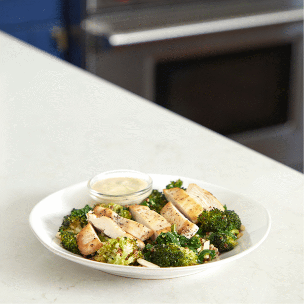 A plate of a ready-to-eat, healthy lunch with Fitlife Foods' Chicken Caesar salad with fiber-rich broccoli, tri-color quinoa, and steamed kale.