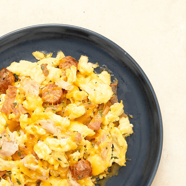A close-up of  gluten-free scrambled eggs, bacon, roasted chicken, sausage, caramelized onions, herbs, & cheddar cheese.