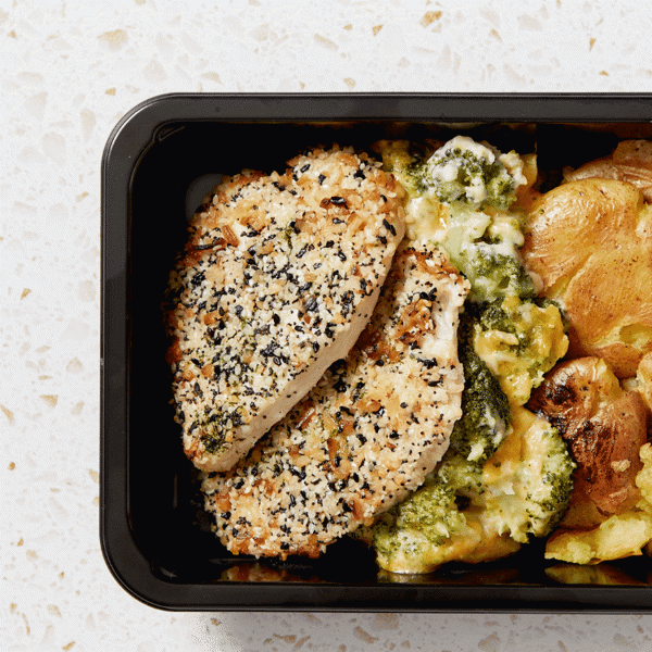 A close-up of pan-seared chicken breast with herbed crust with a side of smashed baby potatoes and cheesy broccoli au gratin in a BPA-free box