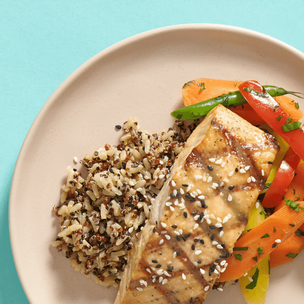 A close up of a freshly prepared dish featuring grilled salmon, miso brushing, herb-roasted veggies, and citrus-soy brown rice quinoa