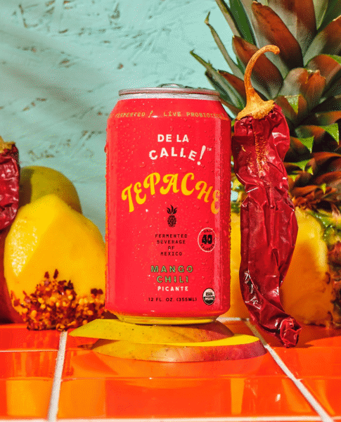 De La Calle Tepache Mango Chili Can with mangoes, chili pepper, and pineapple in the back
